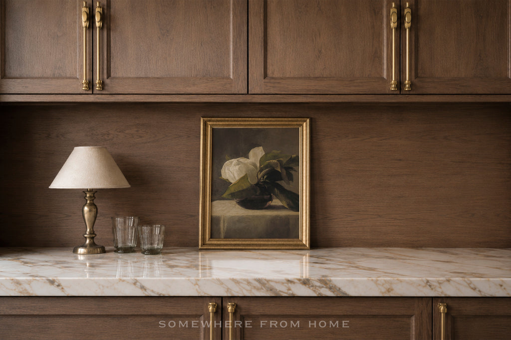 A framed still life painting with white flowers and dark leaves, set in a gold frame on a white marble countertop against a dark wood backsplash. Above, matching dark wood cabinets with gold handles are visible, along with a table lamp and two glasses on the counter.