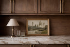 A framed landscape painting with a gold frame sits on a white marble countertop against a dark wood cabinet wall, next to a table lamp and two clear drinking glasses.