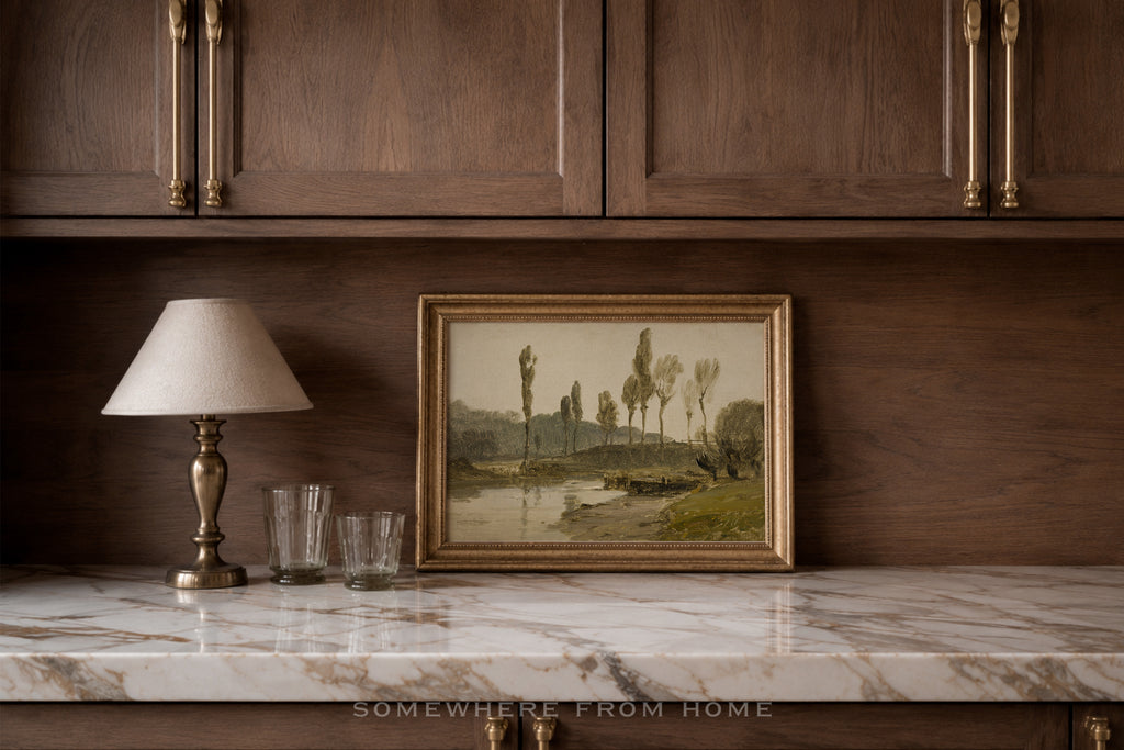 A framed landscape painting with a gold frame sits on a white marble countertop against a dark wood cabinet wall, next to a table lamp and two clear drinking glasses.