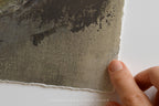 A close-up of a textured handmade paper sheet with a torn edge, held by a person's fingers against a plain white background. The paper has a mottled, organic pattern in gray, brown, and black tones.