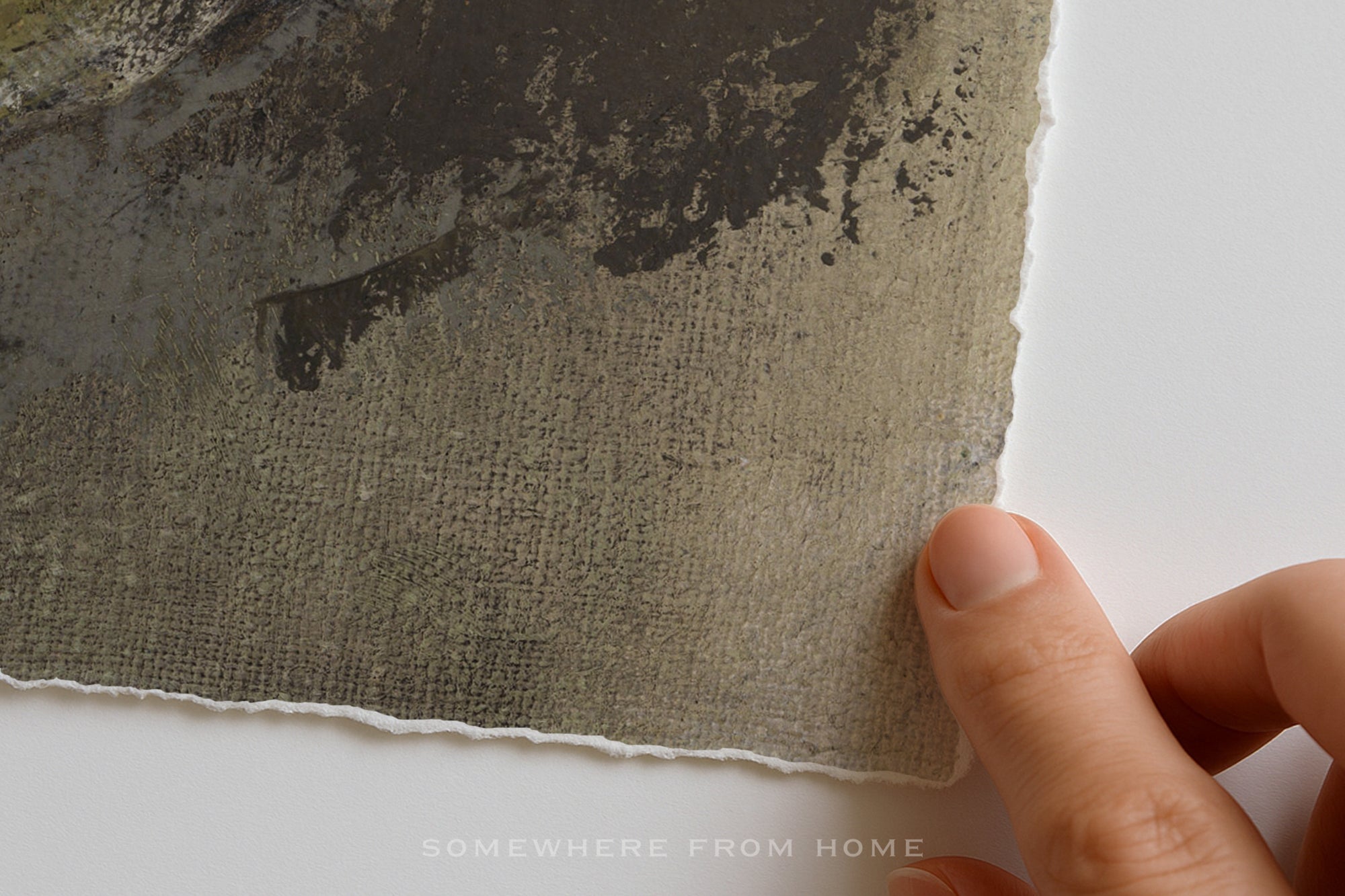 A close-up of a textured handmade paper sheet with a torn edge, held by a person's fingers against a plain white background. The paper has a mottled, organic pattern in gray, brown, and black tones.