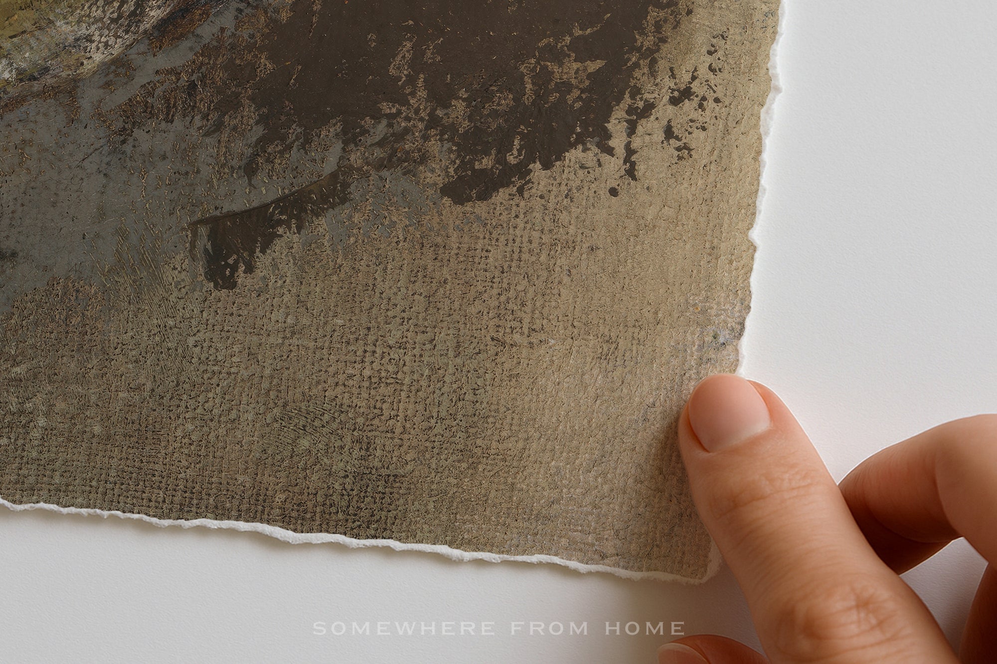 A close-up of a textured handmade paper sheet with a rustic, watercolor-like dark brown pattern on a white background, held by a person's fingers. The watermark 'SOMEWHERE FROM HOME' is visible at the bottom.