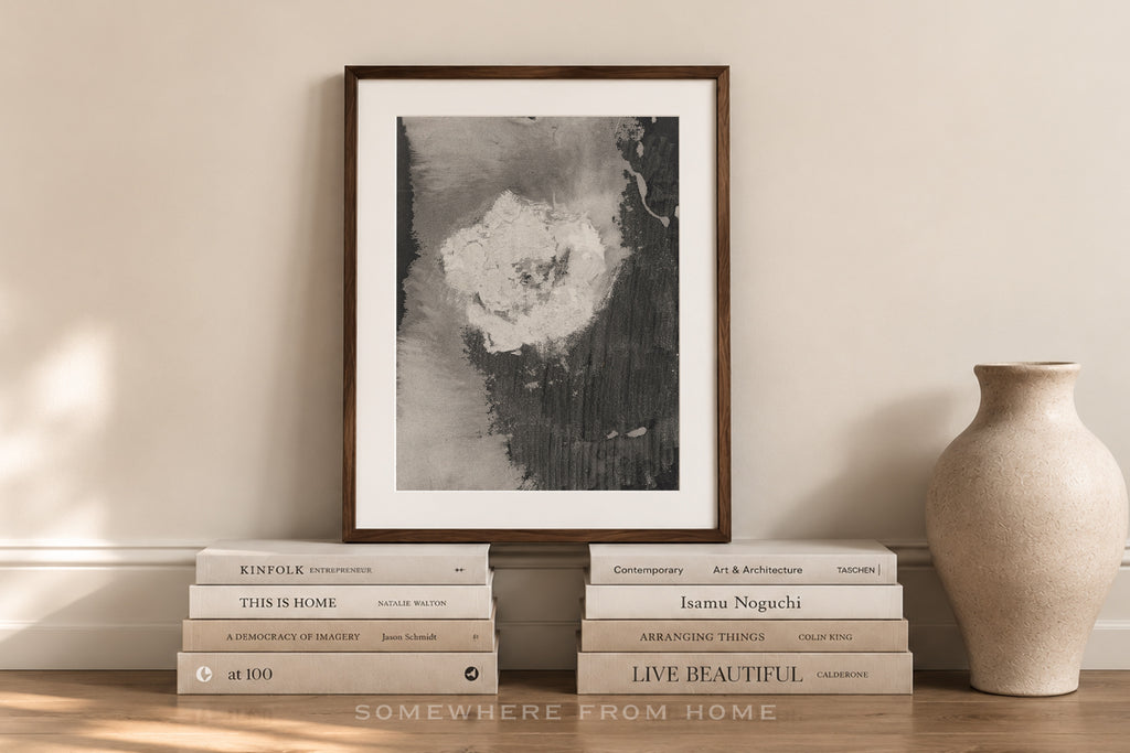 A framed black and white abstract art print with a dark wood frame, styled on a wooden shelf with books and a ceramic vase against a beige wall.