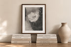 A framed black and white abstract art print with a dark wood frame, styled on a wooden shelf with books and a ceramic vase against a beige wall.