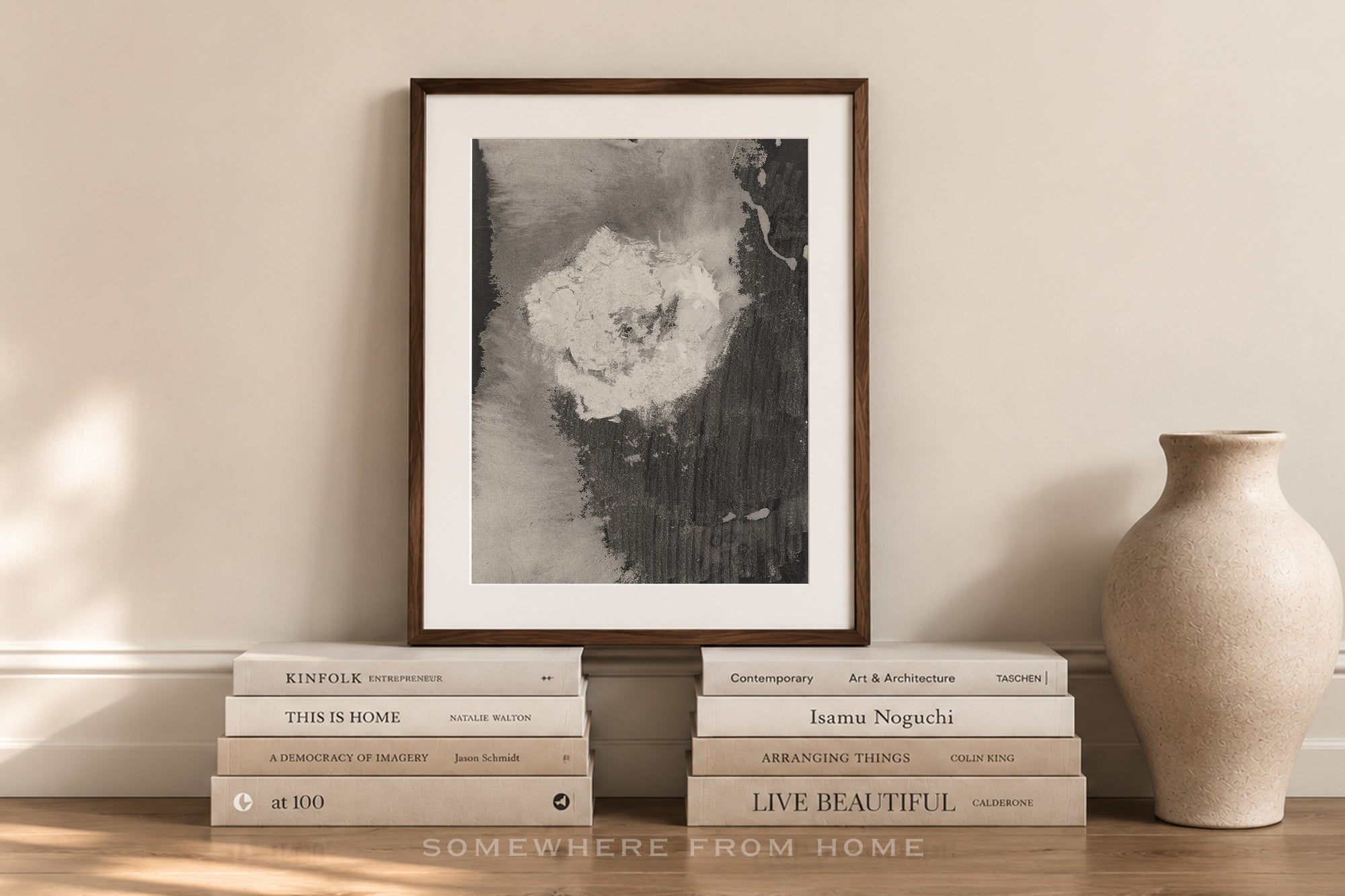 A framed black and white abstract art print with a dark wood frame, styled on a wooden shelf with books and a ceramic vase against a beige wall.