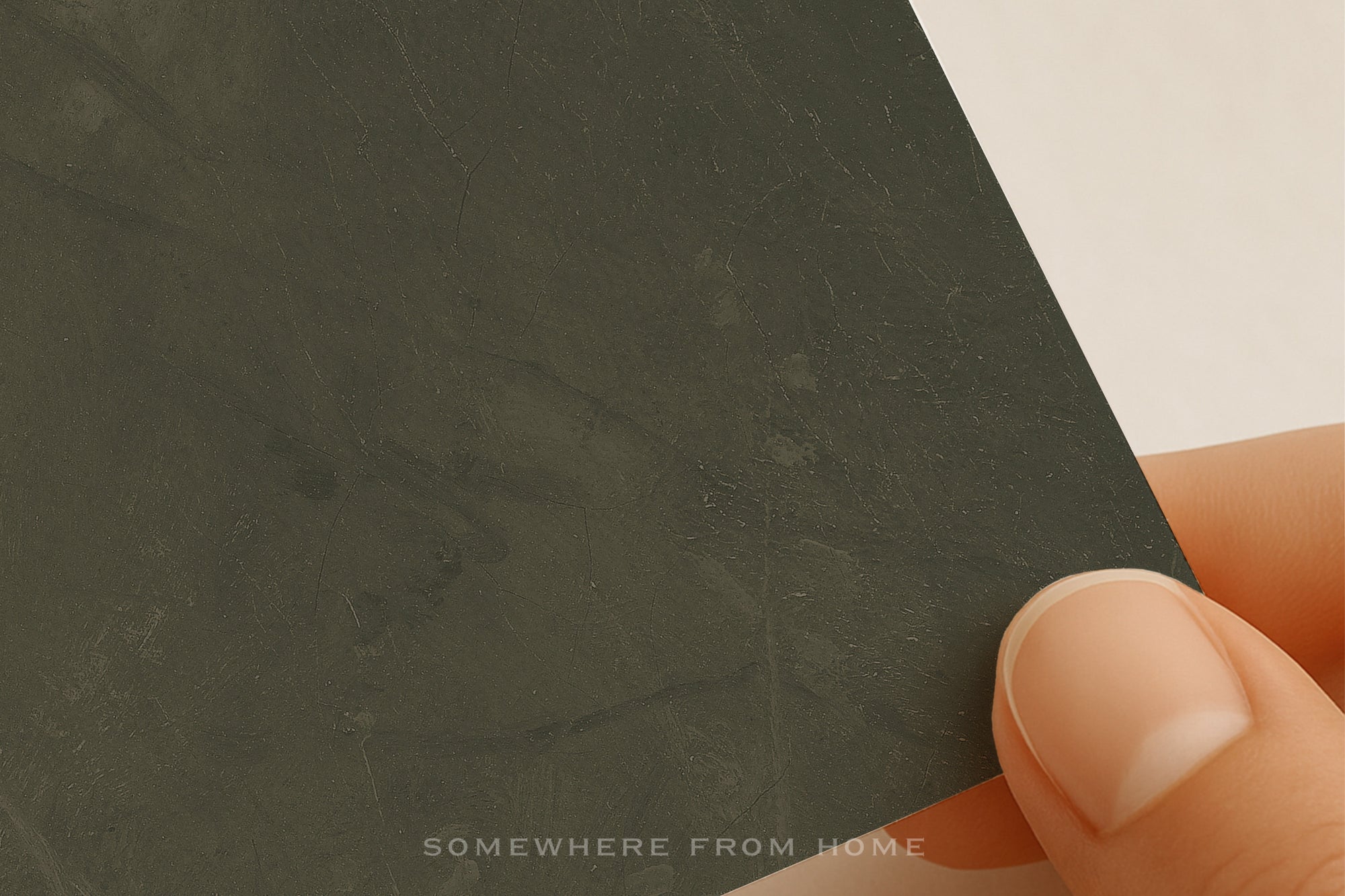 Hand holding a dark green textured art print with 'SOMewhere from home' text.