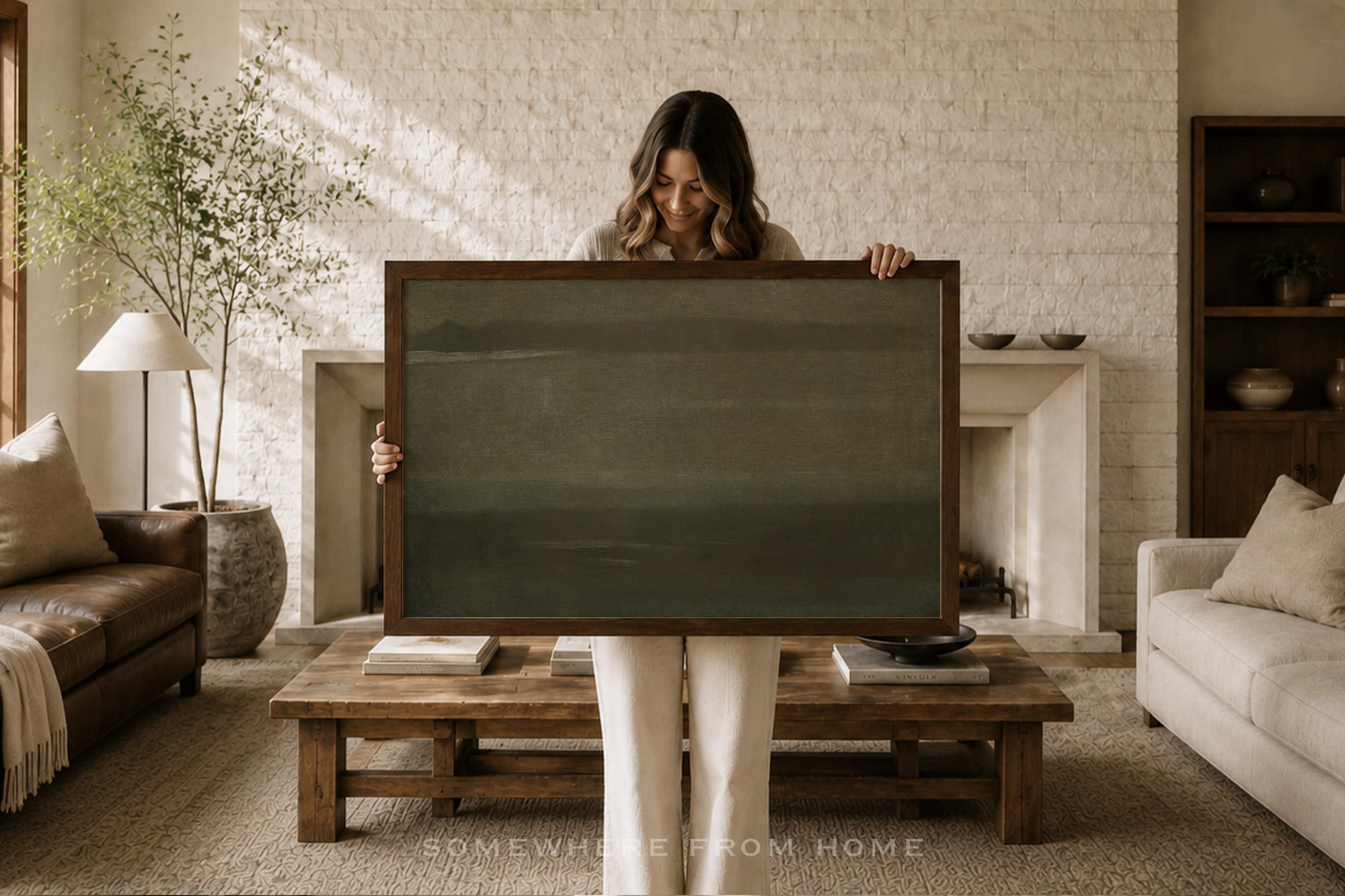 A woman stands in a cozy living room holding a large, dark framed canvas or print. The artwork itself is a muted, dark green landscape.