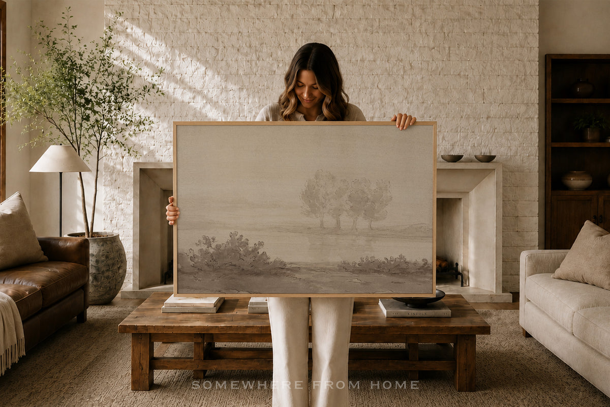 A woman stands in a cozy living room holding up a large framed landscape art print. The artwork features a minimalist, misty tree scene in soft gray and beige tones.