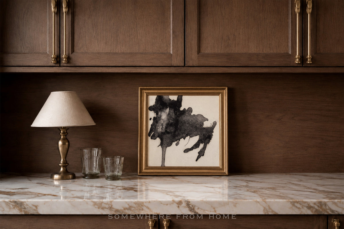 A framed black and white abstract painting with a gold frame sits on a white marble countertop in a kitchen setting with dark wood cabinets and a table lamp.
