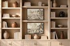 A symmetrical beige built-in bookshelf with two framed sepia landscape prints centered on the middle shelves, surrounded by books, ceramic vases, and bowls.