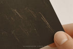 A close-up of a hand holding a dark brown textured art print with faint cream chalk-like scribbles, against a light neutral background. A small 'SOMEWHERE FROM HOME' watermark appears at the bottom.