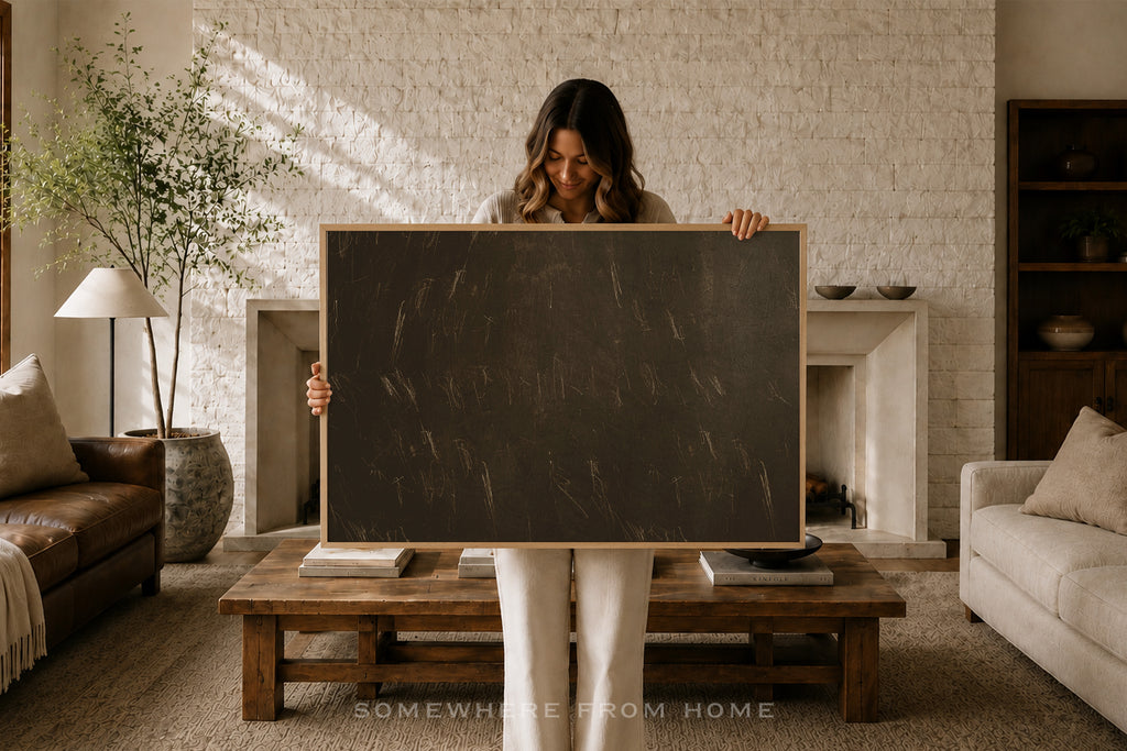 A woman stands in a cozy living room holding a large blank blackboard with a light wood frame. The room features a white brick fireplace, a wooden coffee table, a leather sofa, and a potted plant in the background.