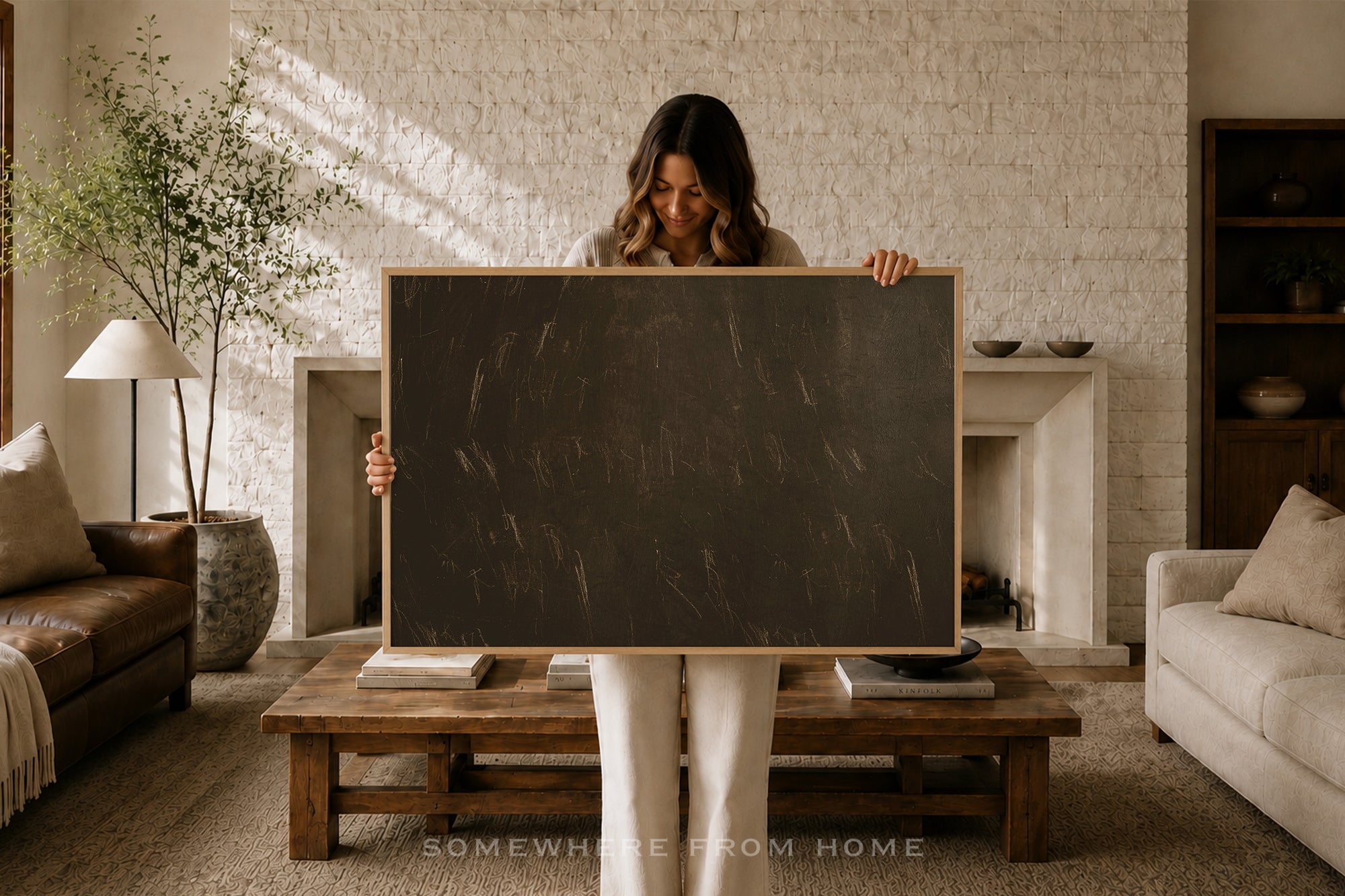 A woman stands in a cozy living room holding a large blank blackboard with a light wood frame. The room features a white brick fireplace, a wooden coffee table, a leather sofa, and a potted plant in the background.