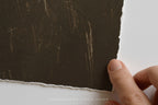 A close-up of a hand holding the edge of a dark brown fine art print with a torn, rustic edge, showing fine lines on the surface, against a white background.