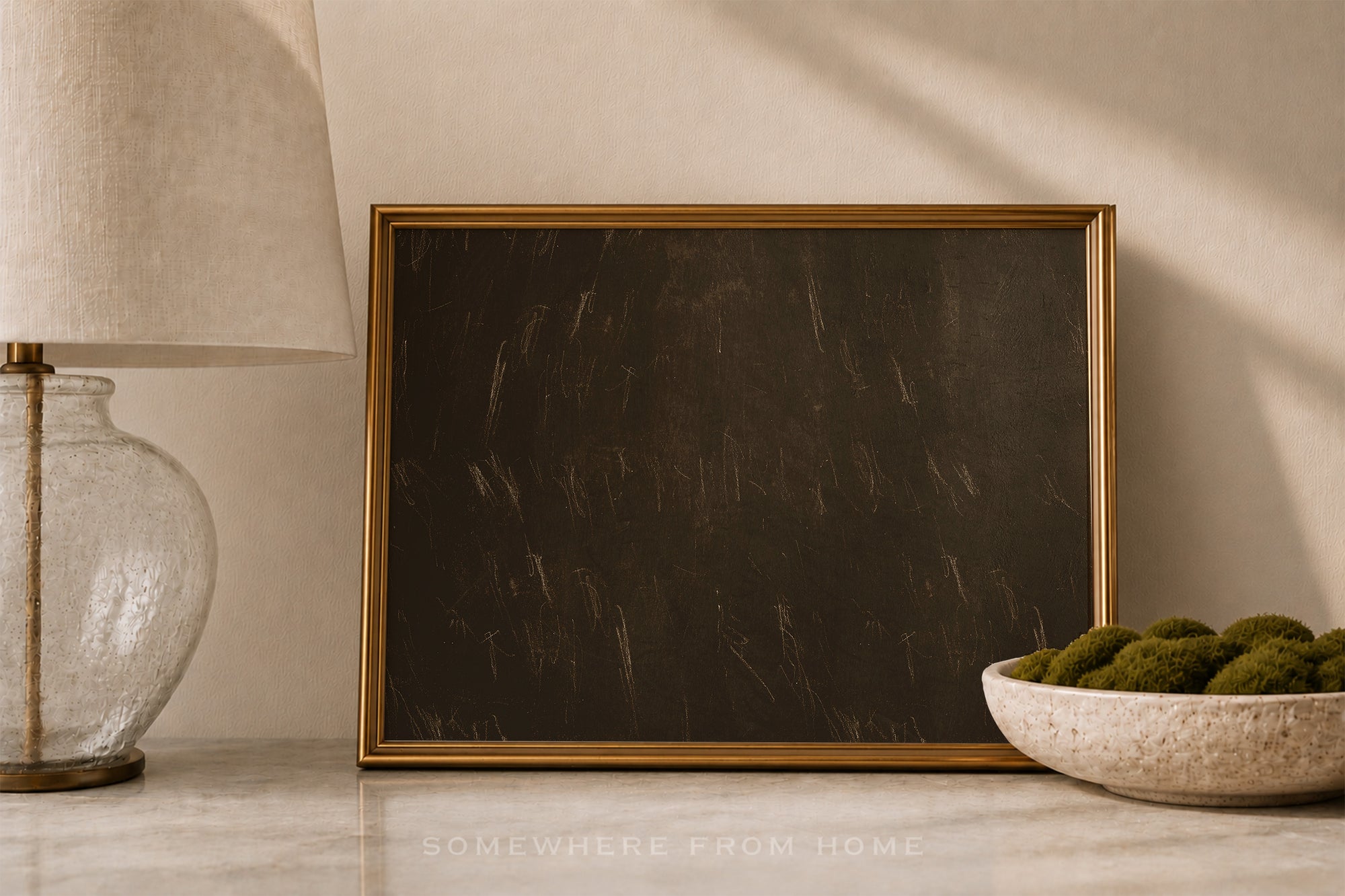 A square framed wall art print with a dark textured surface and a thin gold frame, styled on a marble surface next to a lamp and a bowl of green moss.
