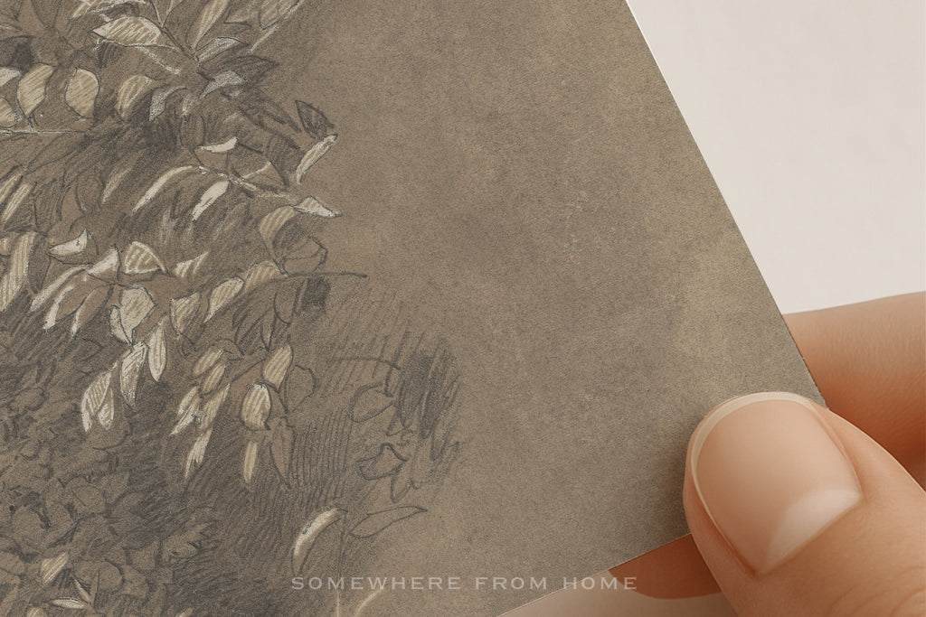 A close-up of a textured gray art print or card being held by a person's fingers, showing a detailed sketch of leaves and branches in gray and white tones.