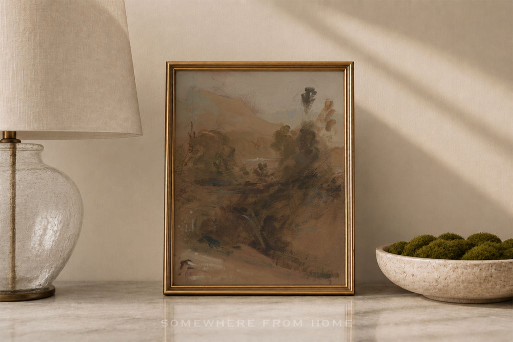 A framed landscape art print with a gold frame, displayed on a marble surface next to a glass table lamp and a bowl of green moss, in a warm, sunlit room.