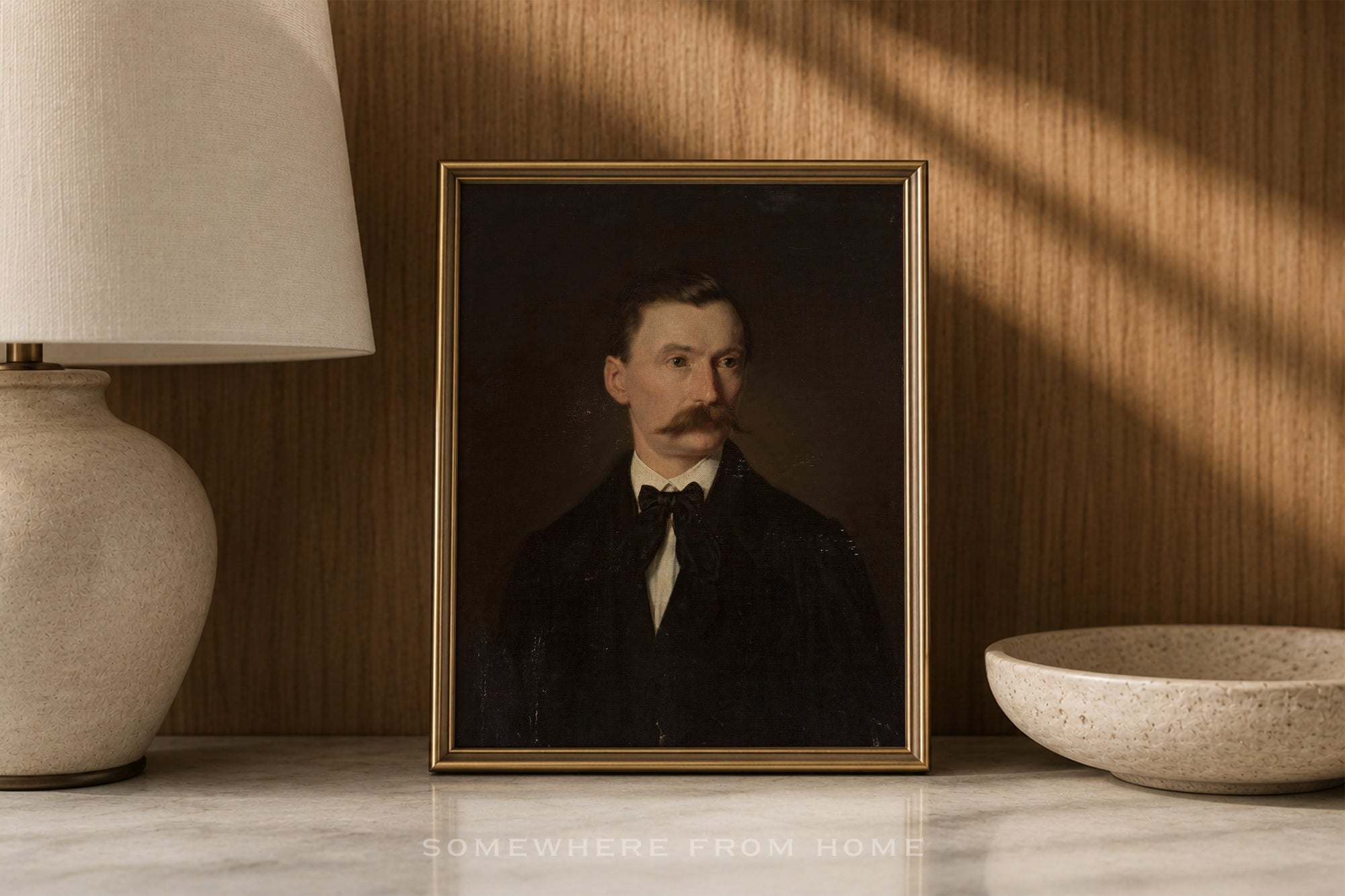 A framed portrait painting of a man with a mustache, displayed on a marble surface against a wood-paneled wall with warm sunlight and shadows. A lamp is visible on the left and a bowl on the right.