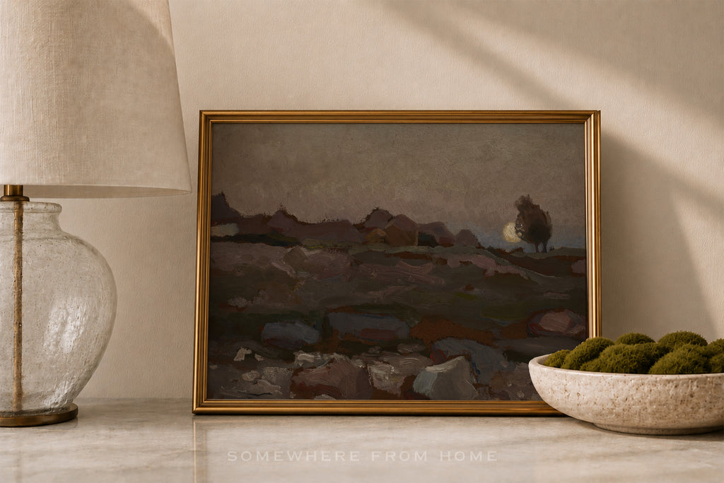 A framed landscape painting with a gold frame, styled on a marble surface next to a table lamp and a bowl of green moss, under warm natural light.