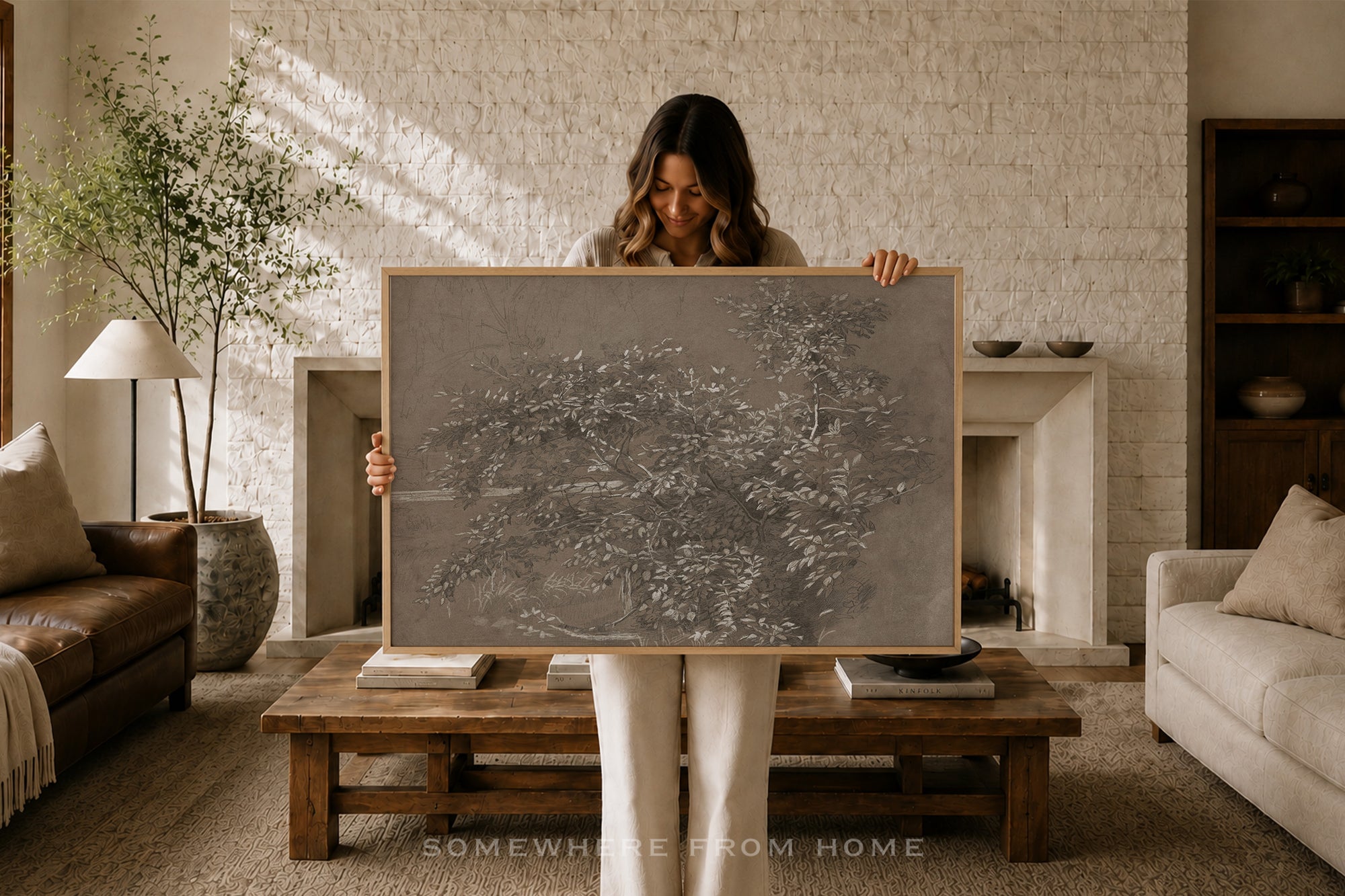 A woman stands in a cozy living room holding up a large framed abstract gray artwork. The room has a brick fireplace, a wooden coffee table, a sofa, and warm sunlight streaming in from the side.