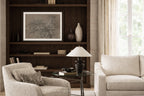 A framed black and white art print displayed on a dark wood built-in bookshelf in a bright, modern living room with beige armchairs and a glass side table.