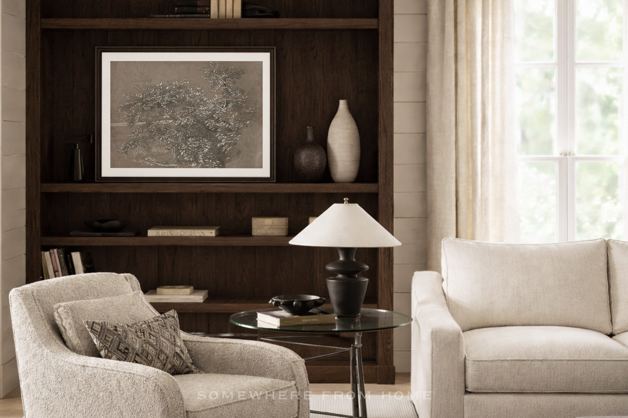 A framed black and white art print displayed on a dark wood built-in bookshelf in a bright, modern living room with beige armchairs and a glass side table.
