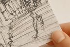A close-up of a hand holding a small square piece of textured paper featuring a black ink sketch of a person walking, with a faint 'FROM HOME' watermark near the bottom.