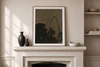 A framed abstract art print with a dark olive-green and black design, displayed on a light beige stone fireplace mantel in a styled room setting.