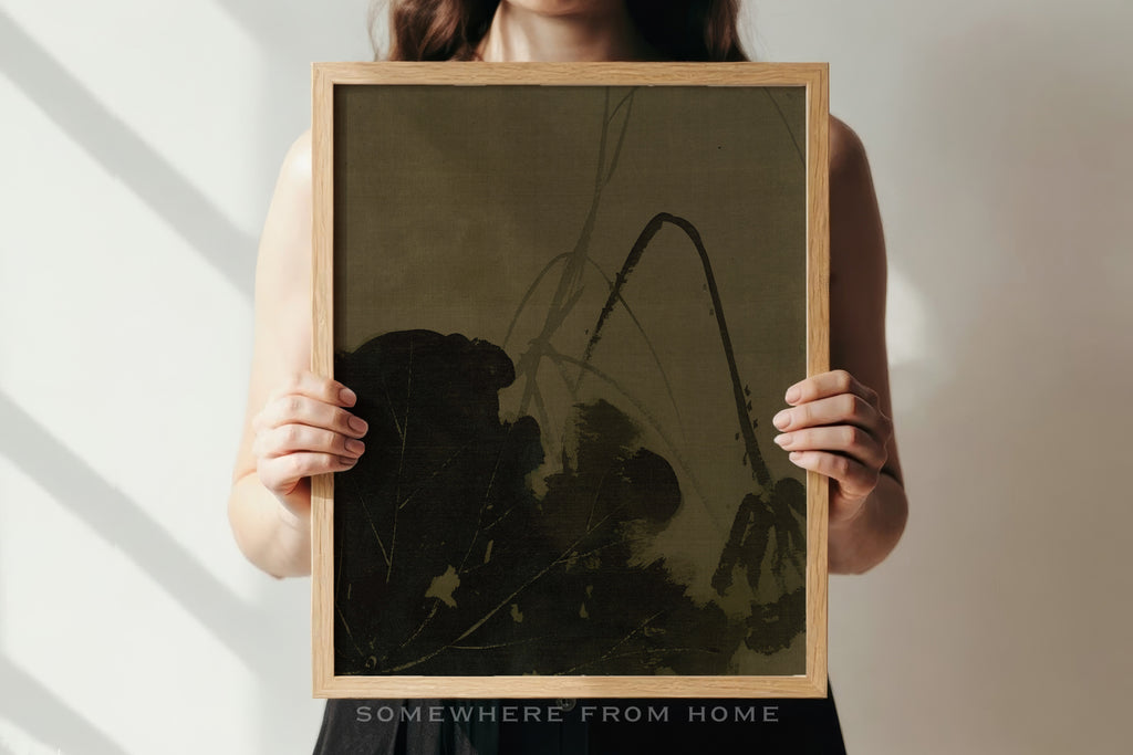 A person holds a square framed art print featuring a dark, abstract botanical design on a muted olive-brown background. The print is enclosed in a light natural wood frame.