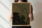 A person holds a square framed art print featuring a dark, abstract botanical design on a muted olive-brown background. The print is enclosed in a light natural wood frame.