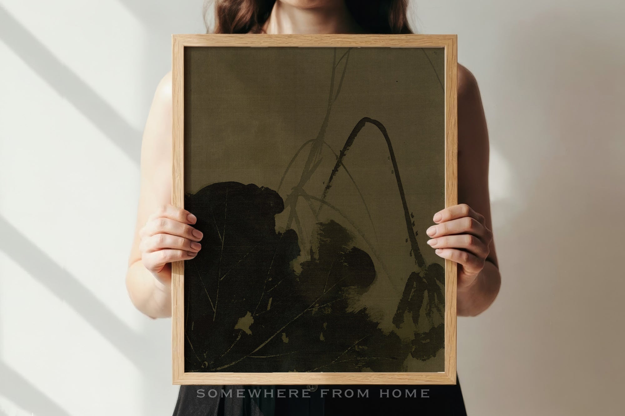 A person holds a square framed art print featuring a dark, abstract botanical design on a muted olive-brown background. The print is enclosed in a light natural wood frame.