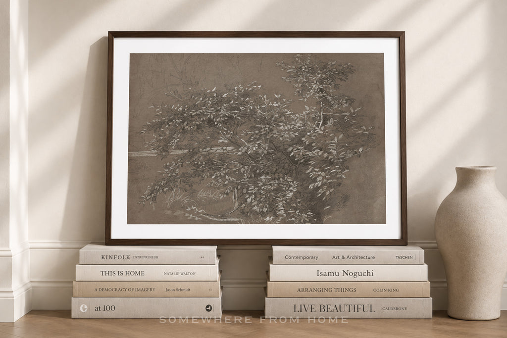A framed monochrome art print leaning against a wall on a wooden floor, styled with stacked books and a ceramic vase in a softly lit interior setting.