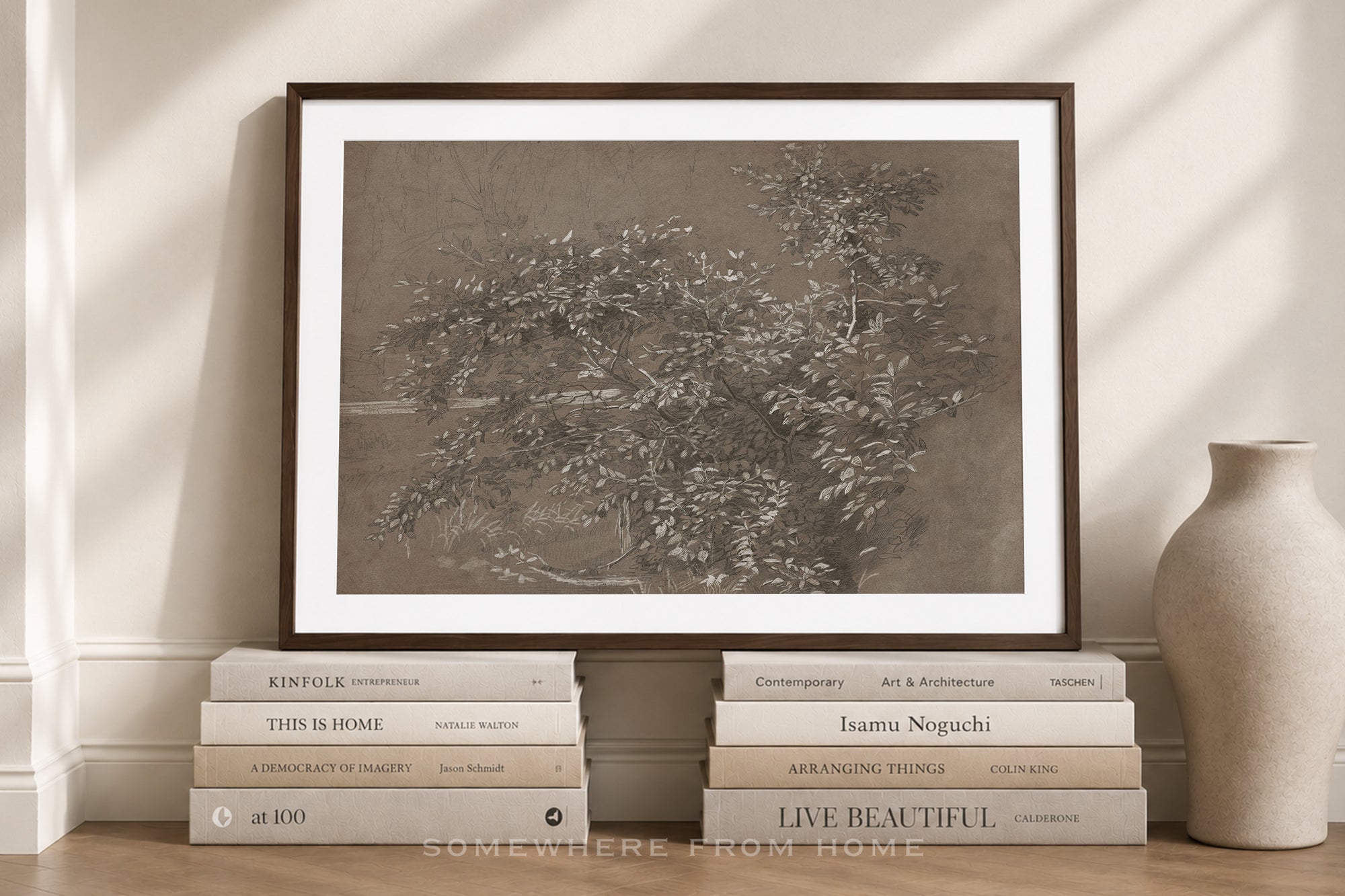 A framed monochrome art print leaning against a wall on a wooden floor, styled with stacked books and a ceramic vase in a softly lit interior setting.