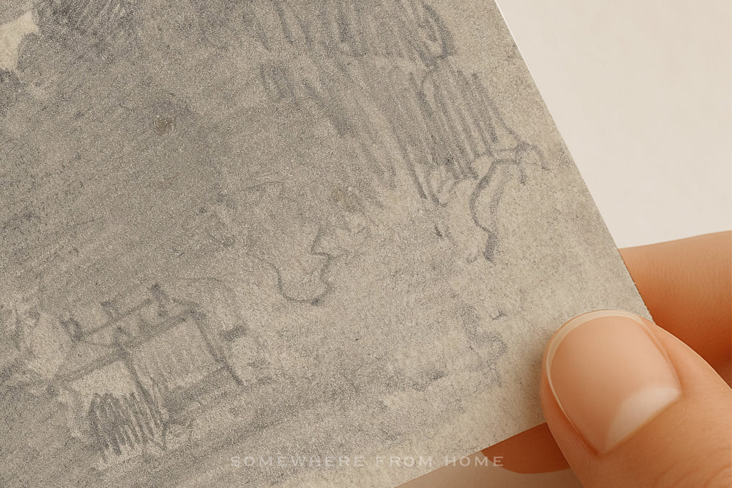 A close-up of a hand holding a square piece of textured gray paper or card with faint pencil-style line drawings, against a plain light background.