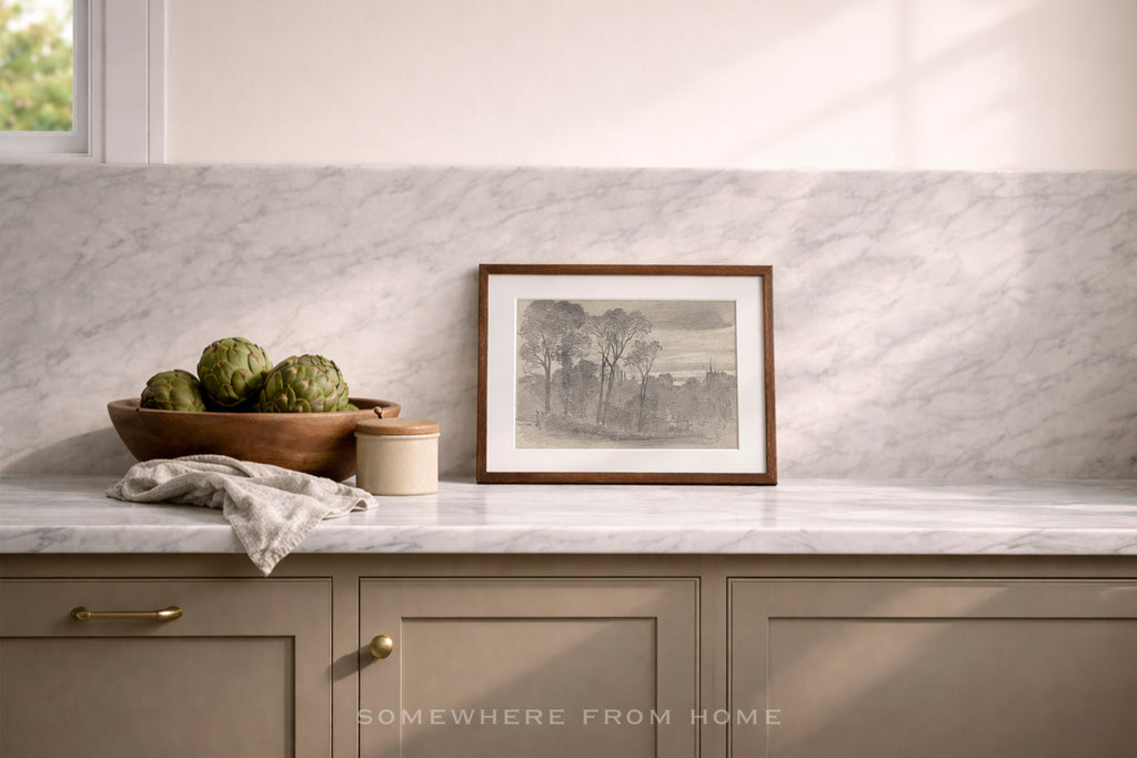 A framed black and white landscape print with a brown wooden frame sits on a white marble kitchen counter against a matching marble backsplash. The scene includes a wooden bowl of artichokes, a small jar, and a folded cloth, with a window light casting soft shadows.