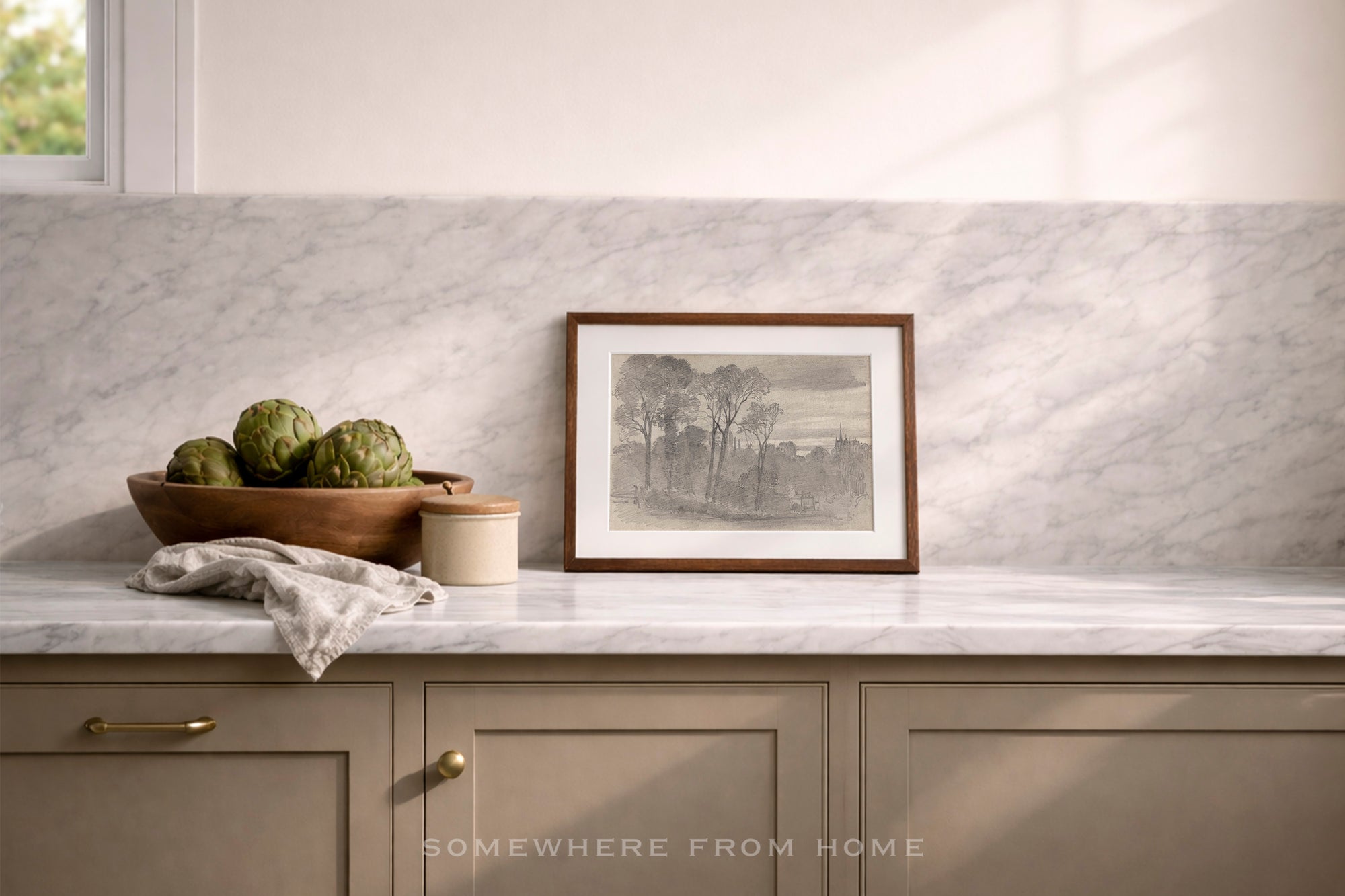 A framed black and white landscape print with a brown wooden frame sits on a white marble kitchen counter against a matching marble backsplash. The scene includes a wooden bowl of artichokes, a small jar, and a folded cloth, with a window light casting soft shadows.