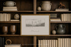 A framed black and white landscape sketch with a gold frame, displayed on a dark wood bookshelf surrounded by books, ceramic pots, and decorative spheres.