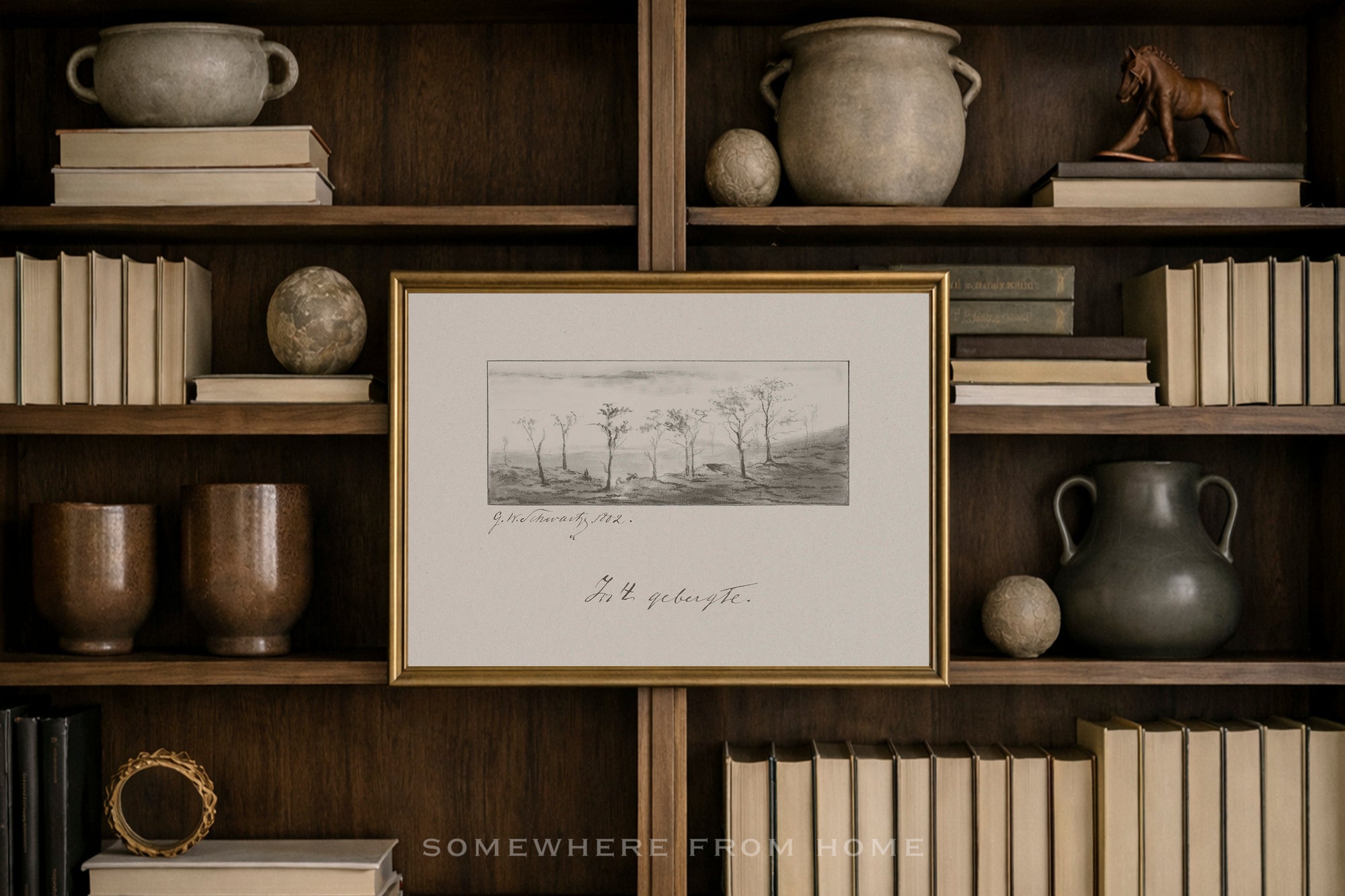A framed black and white landscape sketch with a gold frame, displayed on a dark wood bookshelf surrounded by books, ceramic pots, and decorative spheres.