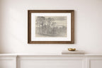 A framed landscape art print with a brown wood frame and white mat, hanging on a white wall above a white console table with a small gold bowl and books.