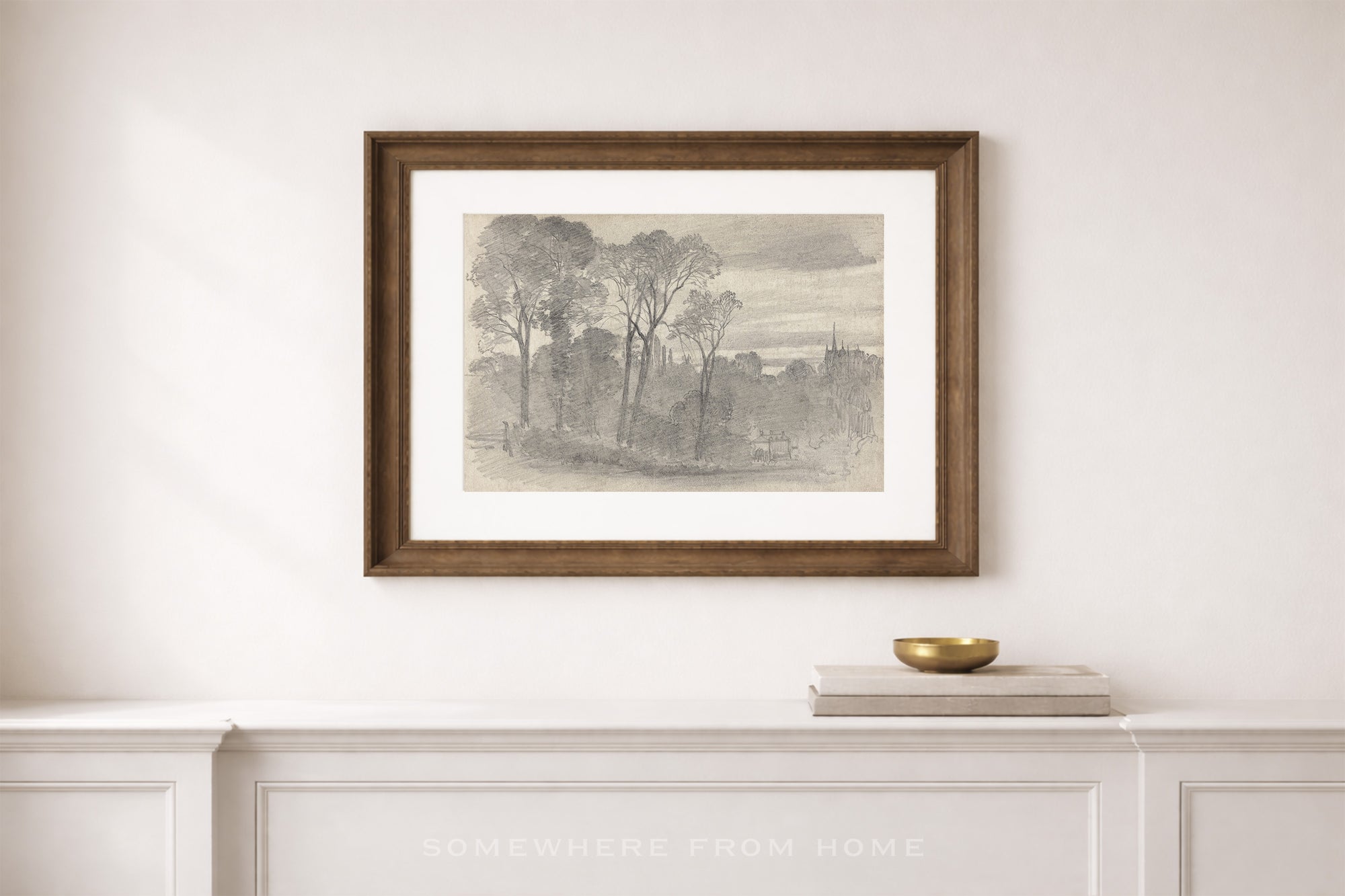 A framed landscape art print with a brown wood frame and white mat, hanging on a white wall above a white console table with a small gold bowl and books.