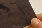 A close-up of a hand holding a textured dark brown art print or card against a plain light background. The print shows a partially visible dark painted design, and a small watermark reading 'SOMEWHERE FROM HOME' appears near the bottom.