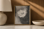 A framed abstract art print with a gold frame, featuring textured gray, black, and white brushstrokes, displayed on a marble surface against a wood-paneled wall with warm sunlight and shadows. A beige ceramic lamp is on the left and a matching bowl is on the right.