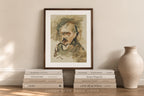 A framed watercolor portrait print of Albert Einstein leaning against a beige wall, styled on a wooden surface with stacked books and a ceramic vase.