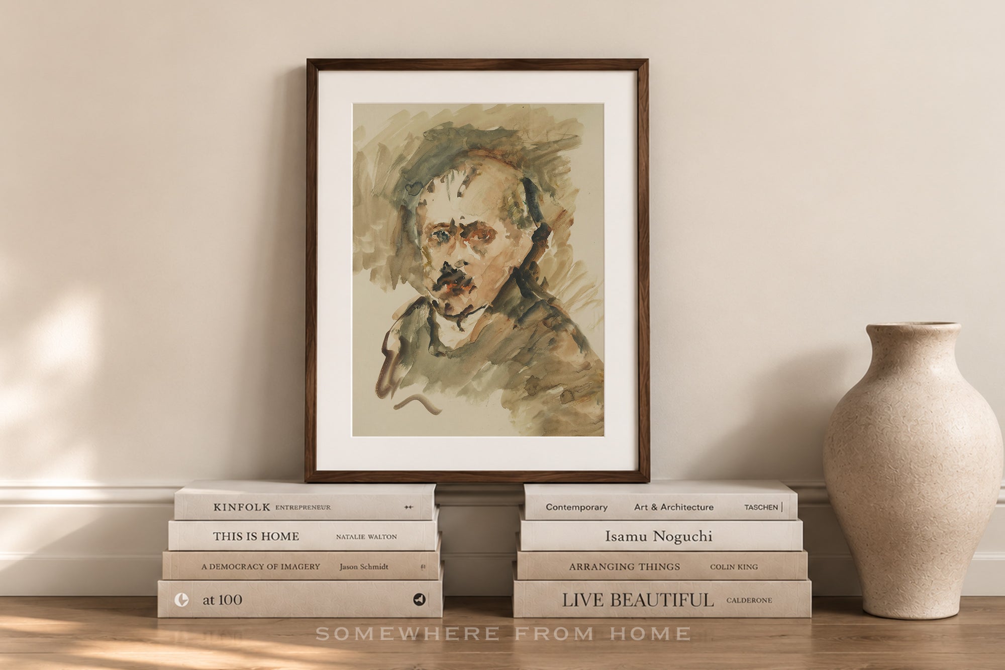A framed watercolor portrait print of Albert Einstein leaning against a beige wall, styled on a wooden surface with stacked books and a ceramic vase.