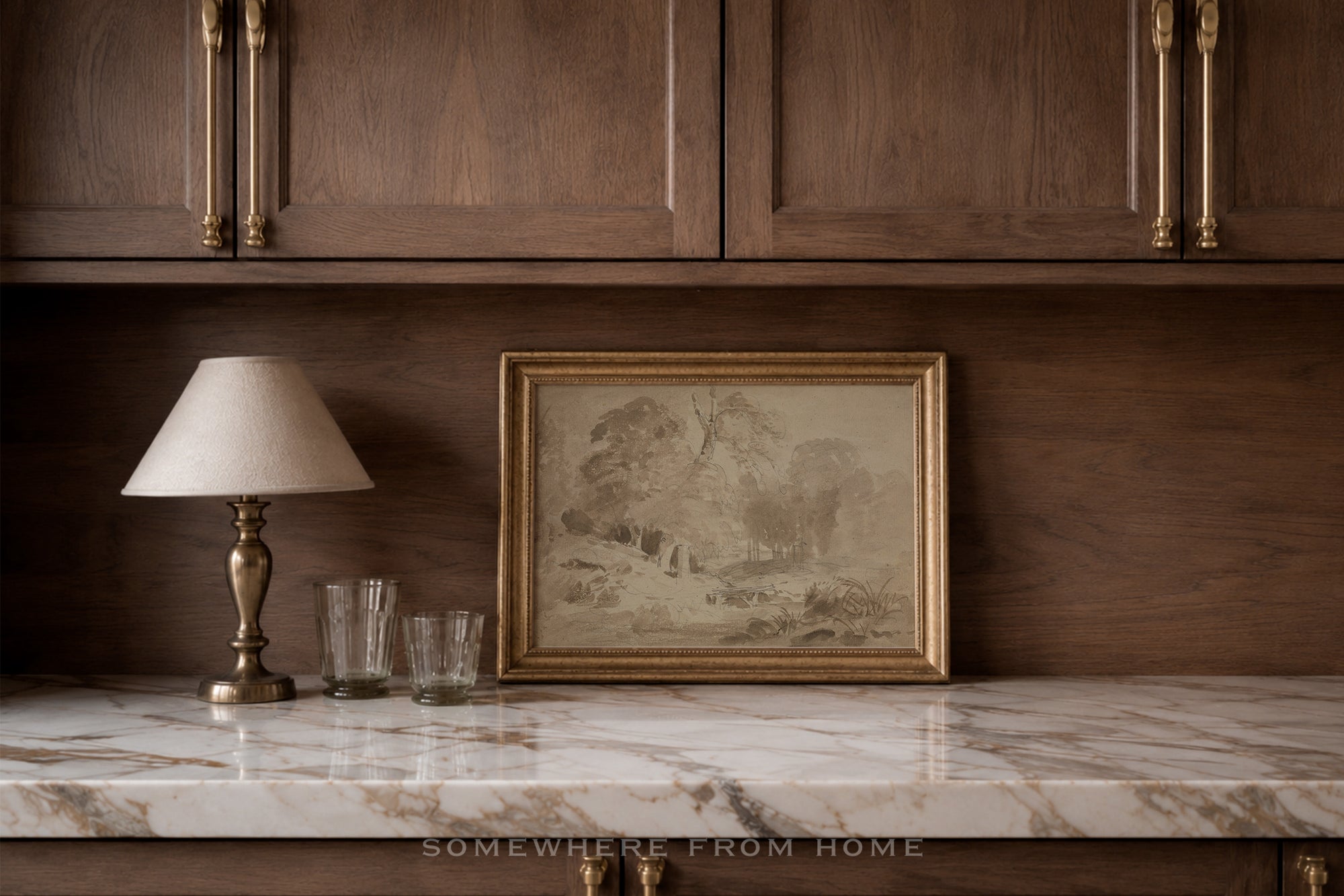 A framed landscape art print with a gold frame sits on a white marble countertop against a dark wood cabinet wall, next to a table lamp and two glasses.