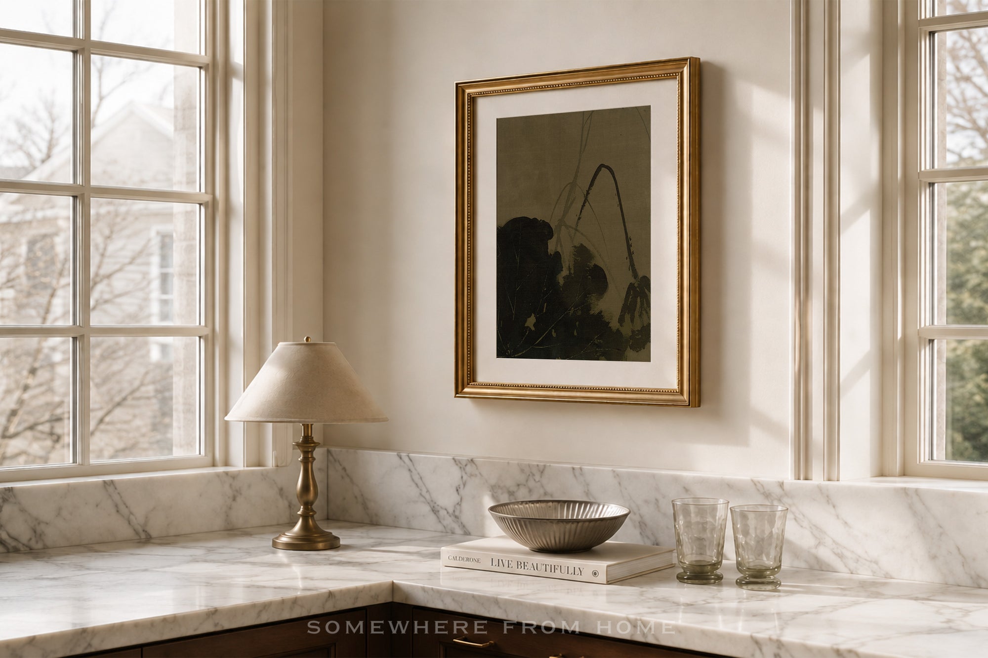A framed abstract art print with a gold frame and white mat hangs on a cream wall above a marble kitchen counter with a lamp, bowl, and glasses.