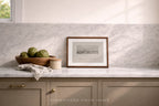 A framed black and white landscape print with a medium brown wooden frame sits on a marble countertop against a white marble backsplash. The scene includes a wooden bowl of artichokes, a small jar, and a folded cloth, with a window light casting soft shadows.