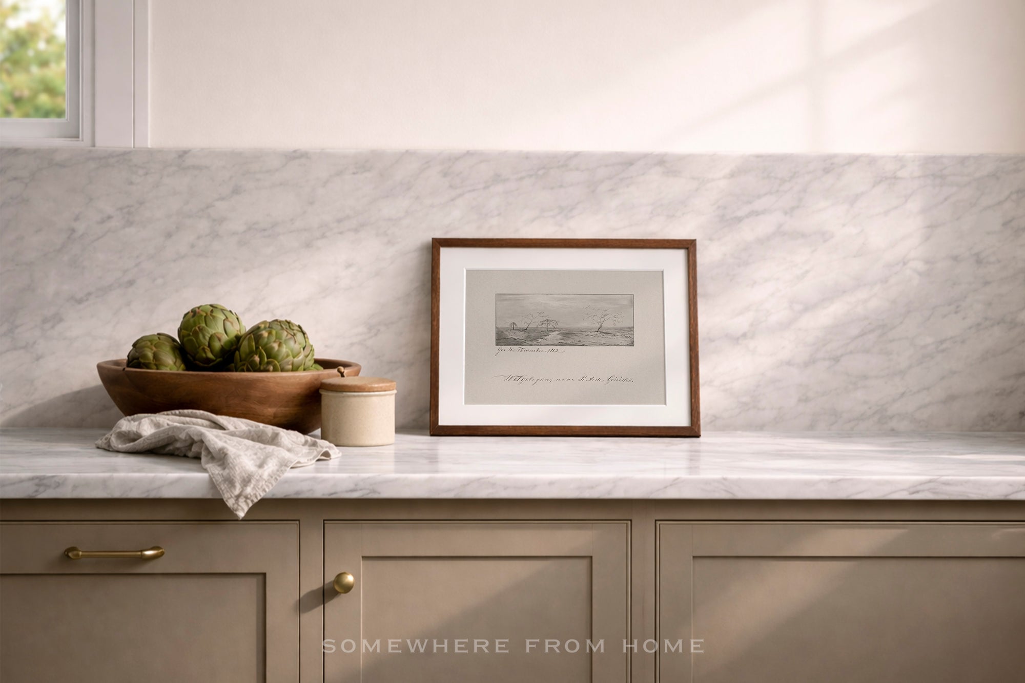 A framed black and white landscape print with a medium brown wooden frame sits on a marble countertop against a white marble backsplash. The scene includes a wooden bowl of artichokes, a small jar, and a folded cloth, with a window light casting soft shadows.