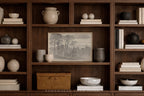 A framed monochrome landscape print displayed on a dark wood bookshelf, surrounded by books, ceramic vases, bowls, and a wicker storage box in a warm, styled interior setting.