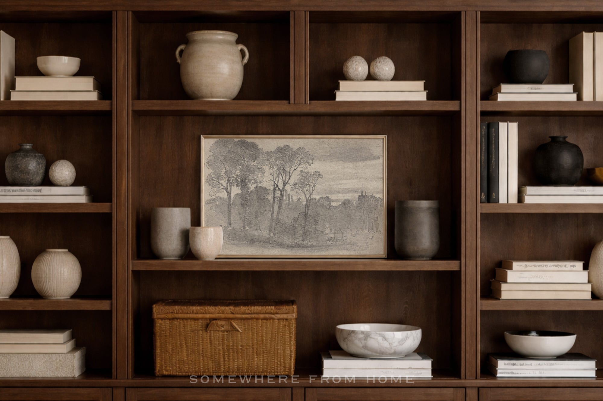 A framed monochrome landscape print displayed on a dark wood bookshelf, surrounded by books, ceramic vases, bowls, and a wicker storage box in a warm, styled interior setting.